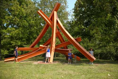 Children play around a large, abstract orange sculpture in a grassy park surrounded by trees.