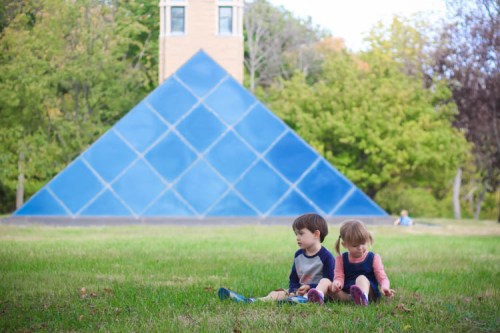Two children sit on grass in front of a blue pyramid structure, surrounded by trees in a park setting.