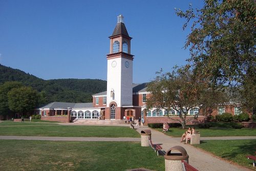 A brick building with a clock tower, surrounded by green grass and trees, under a clear blue sky. People are walking nearby.