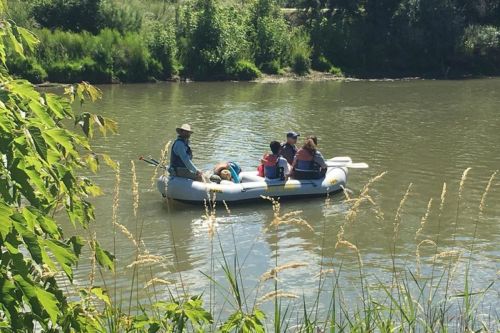 A group of five people in a raft on a calm river, surrounded by greenery and tall grass.
