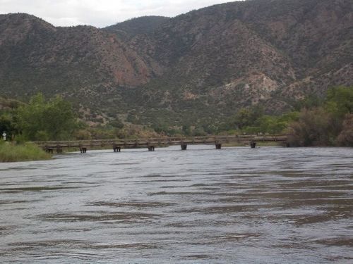 A serene river flows through a mountainous landscape, with a wooden bridge crossing in the distance.