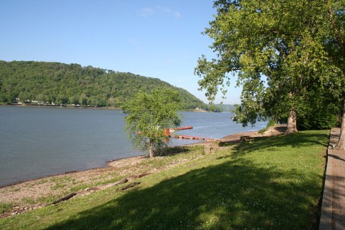 A serene riverbank scene with green trees, a dock, and a gentle slope leading to the water under a clear blue sky.