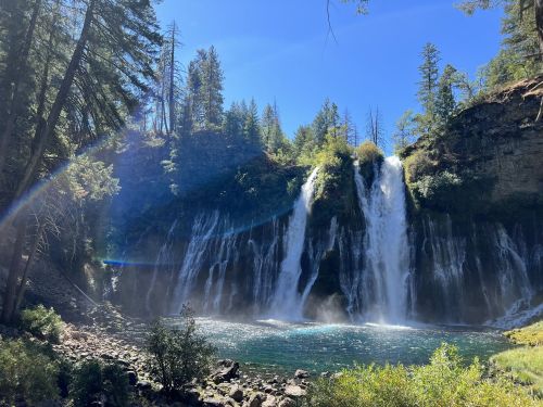 A scenic waterfall cascades into a clear pool, surrounded by lush trees and a bright blue sky.