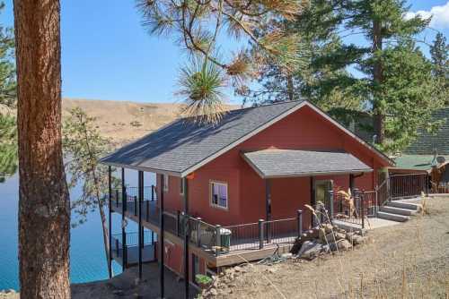 A red lakeside cabin surrounded by trees, with a deck overlooking the water and hills in the background.