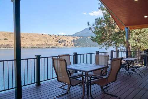 A scenic lakeside view from a deck with tables and chairs, surrounded by mountains and clear blue skies.