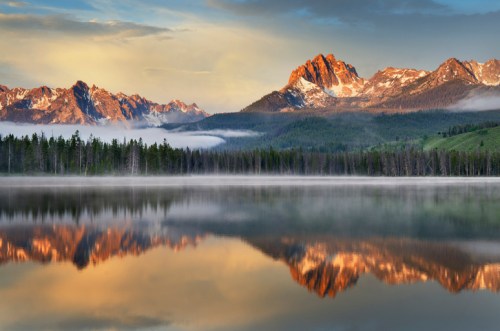 A serene lake reflects majestic mountains and a colorful sky at sunrise, surrounded by lush forests.