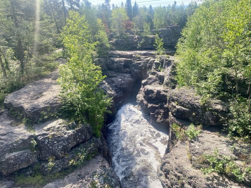 A rocky landscape with a flowing river surrounded by lush green trees and sunlight filtering through the foliage.