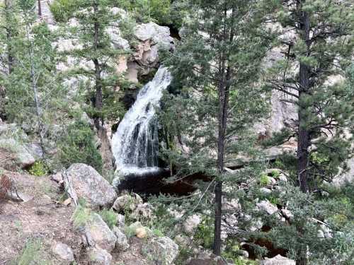 A serene waterfall cascades down rocky terrain, surrounded by lush green trees and boulders.