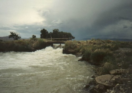 A turbulent river flows under a small bridge, surrounded by lush greenery and dark storm clouds in the sky.