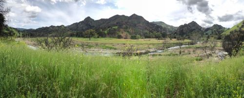 A panoramic view of green fields and mountains under a partly cloudy sky, with a winding river in the foreground.