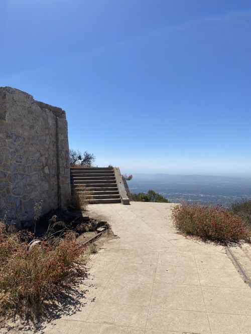 A stone structure beside a pathway leading to stairs, with a clear blue sky and distant hills in the background.