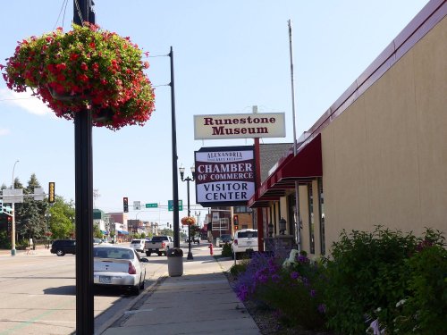 A street view featuring the Runestone Museum and Chamber of Commerce Visitor Center, with hanging flower baskets.