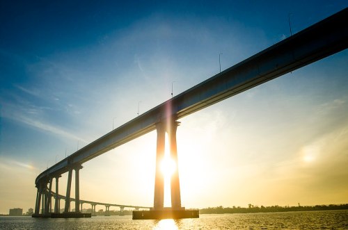 Silhouette of a bridge over water at sunset, with sunlight shining through the structure and a clear blue sky.