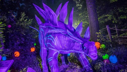 A large, purple dinosaur sculpture with spikes, illuminated in a dark forest, surrounded by colorful pumpkins.
