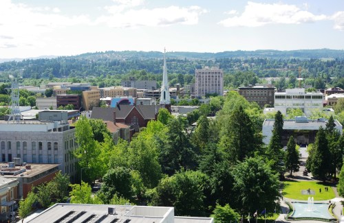 A panoramic view of a cityscape featuring buildings, trees, and a church steeple under a clear blue sky.