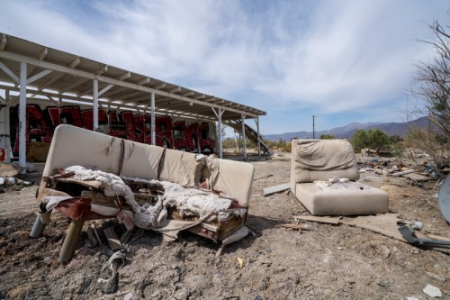 Abandoned, worn-out furniture sits in a desolate area with debris and a building in the background under a cloudy sky.