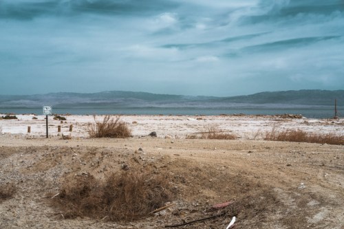 A barren landscape with dry earth, sparse vegetation, and a distant view of mountains under a cloudy sky.