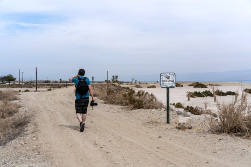 A person walks along a dirt path near a "No Trespassing" sign, with dry vegetation and a cloudy sky in the background.