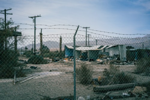 A desolate landscape with abandoned buildings, overgrown vegetation, and a barbed wire fence under a cloudy sky.