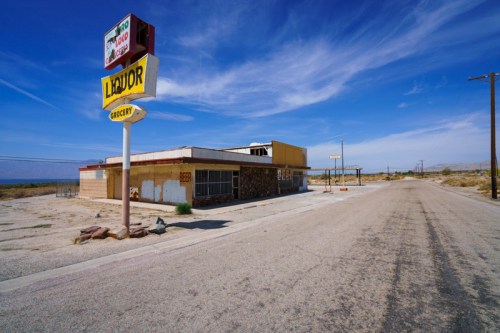Abandoned grocery store with a liquor sign, set against a clear blue sky and empty road in a desolate area.
