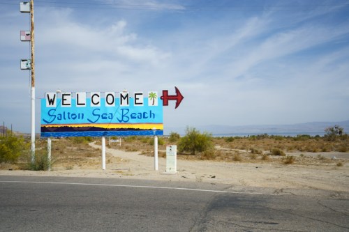A colorful welcome sign for Salton Sea Beach, with a desert landscape and blue sky in the background.