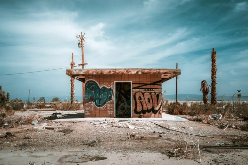 Abandoned building with graffiti, surrounded by dry vegetation and palm trees under a cloudy sky.