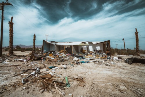 Abandoned building surrounded by debris and trash, with a cloudy sky and barren landscape in the background.