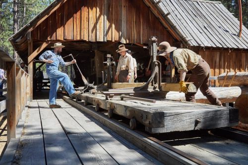 Three men in vintage clothing operate a sawmill, cutting logs in front of a rustic wooden building in a forested area.