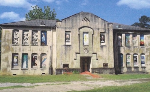 Abandoned building with weathered walls and colorful murals on windows, set against a blue sky and green grass.