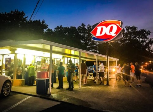 A crowd of people waiting in line outside a Dairy Queen at dusk, illuminated by the restaurant's bright sign.