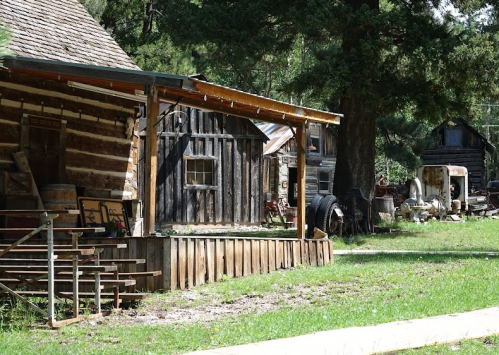 A rustic wooden cabin with a porch, surrounded by greenery and old structures in a serene outdoor setting.