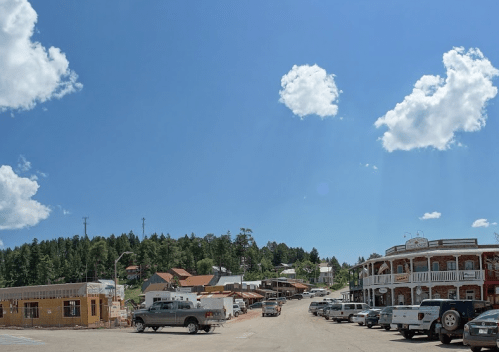 A sunny street scene in a small town, featuring shops, parked cars, and trees under a blue sky with fluffy clouds.