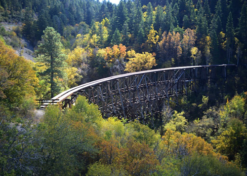 A wooden trestle bridge arches over a forested valley filled with autumn foliage.