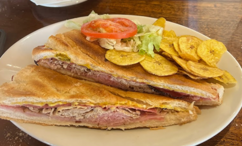 A Cuban sandwich with layers of meat, pickles, and mustard, served with lettuce, tomato, and plantain chips on the side.