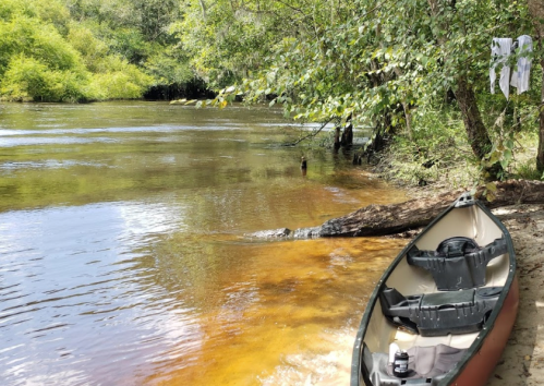 A canoe by a calm riverbank surrounded by trees, with clothes hanging and bags on the sandy shore.