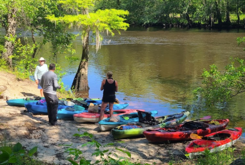 Three people by a river, preparing kayaks on the shore surrounded by trees and greenery.