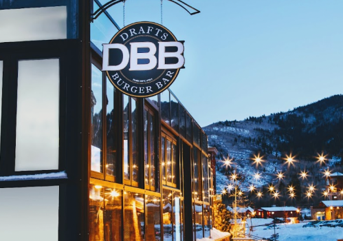 Sign for "DBB Drafts Burger Bar" outside a modern building, with a snowy mountain backdrop and twinkling lights.