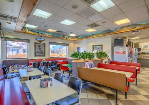 A cozy diner interior with red and beige seating, tables, and decor, featuring plants and a Coca-Cola fridge.