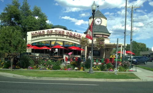 A restaurant with a clock tower, outdoor seating, and colorful flowers, under a blue sky with scattered clouds.