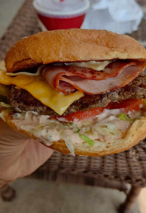 A close-up of a loaded burger with cheese, ham, pickles, lettuce, tomato, and sauce, held in a hand.