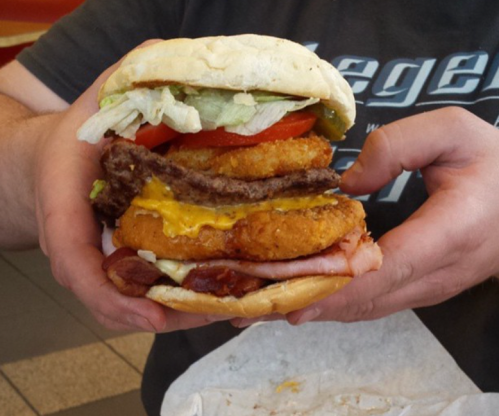 A person holds a large, stacked burger with lettuce, tomato, cheese, chicken patty, and bacon, showcasing its size.