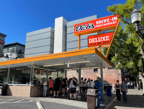 A busy drive-in restaurant with a bright sign, featuring a crowd of people waiting in line outside.