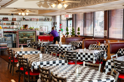 A diner interior with checkered tablecloths, red booths, and a counter, featuring two staff members and a cozy atmosphere.