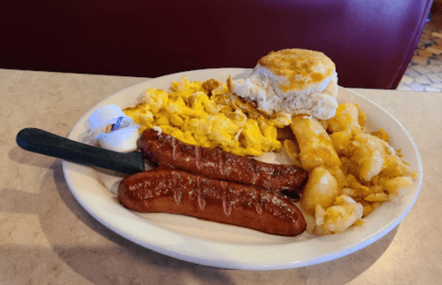 A plate of breakfast featuring scrambled eggs, two sausage links, hash browns, a biscuit, and a butter packet.