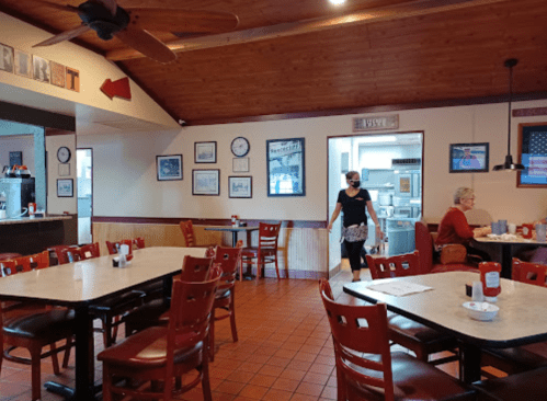 A cozy diner interior with wooden accents, tables, and a woman walking past a booth. Wall clocks and framed photos decorate the space.