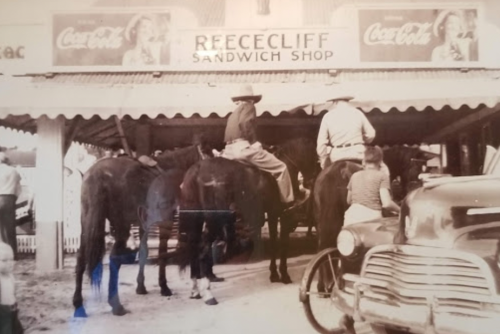 Two cowboys on horseback in front of a vintage sandwich shop, with an old car parked nearby. Black and white photo.
