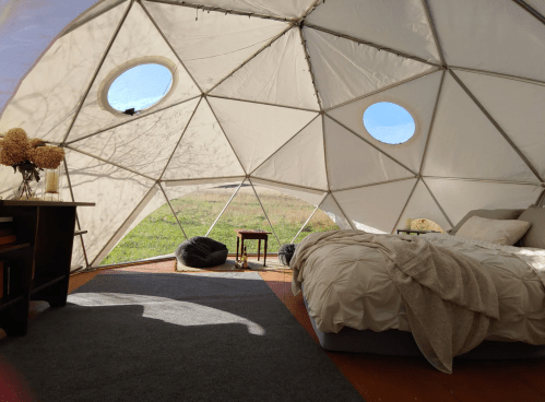 Interior of a geodesic dome with a bed, cozy decor, and large circular windows showing a grassy landscape outside.