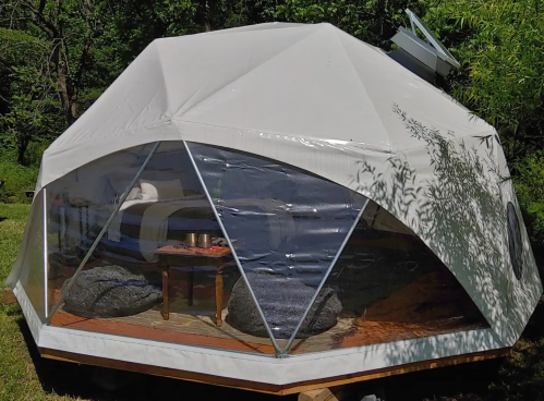 A geodesic dome tent with large windows, set in a green outdoor area, featuring a cozy interior with bean bags and a table.