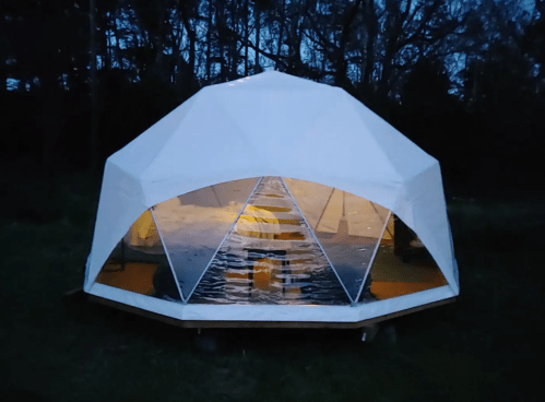 A geodesic dome structure illuminated at dusk, surrounded by trees and grass.