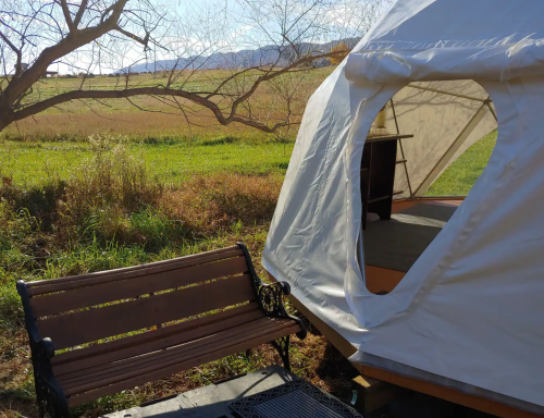 A cozy tent beside a wooden bench, set against a backdrop of green fields and distant mountains under a clear blue sky.